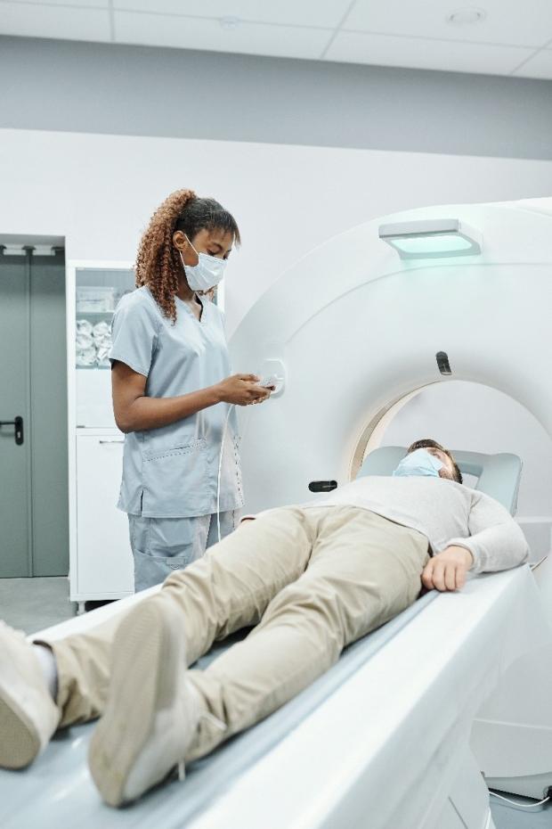 A nurse wearing a face mask talking to a person lying on a couch next to a donut-shaped scanner machine.