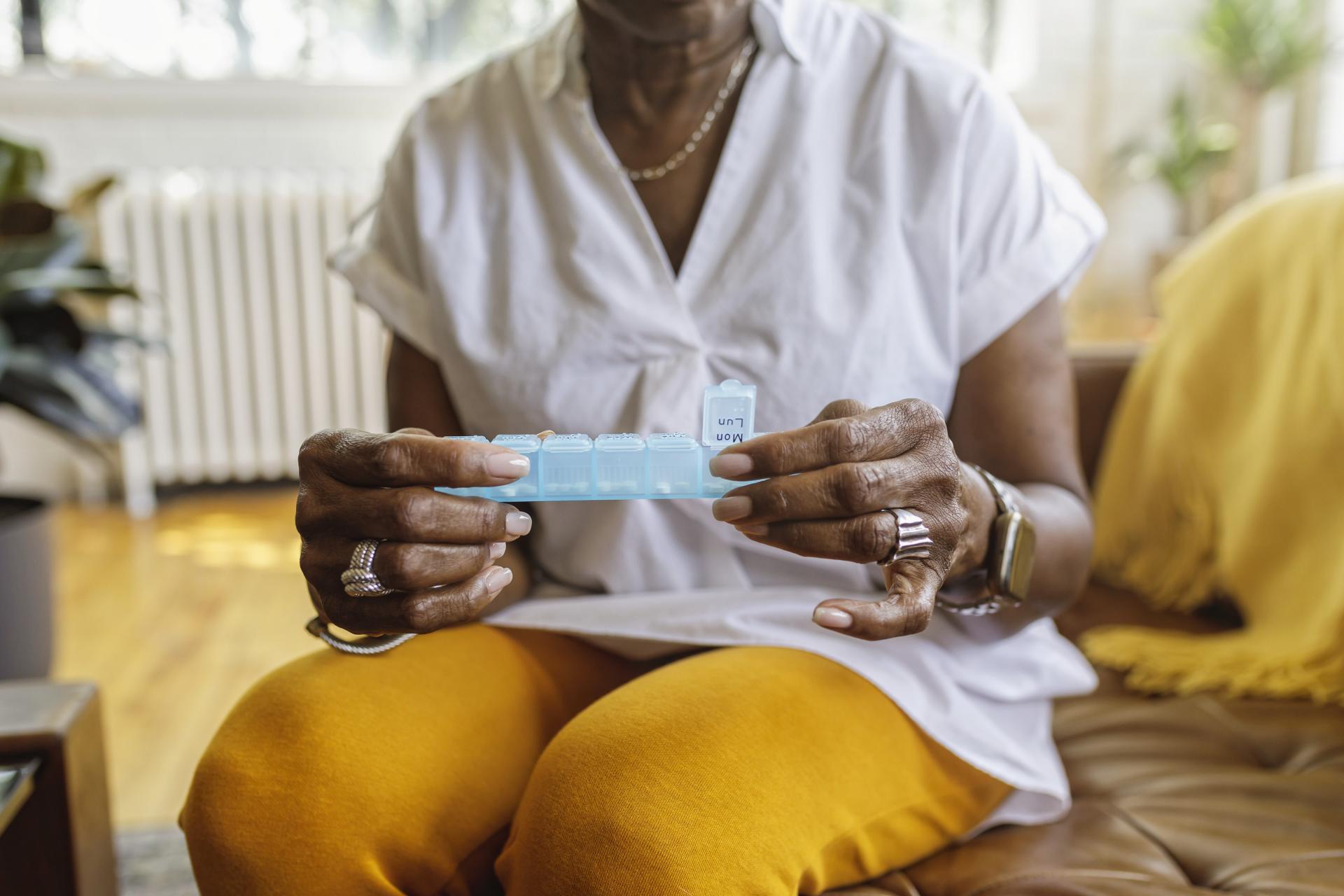 Person in a t-shirt and trousers sits on a sofa. In their hands is an open pill box medication aid.