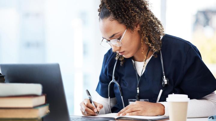 A healthcare professional sitting at a desk studying, writing in a notebook with a laptop in front and a cup and pile of books 