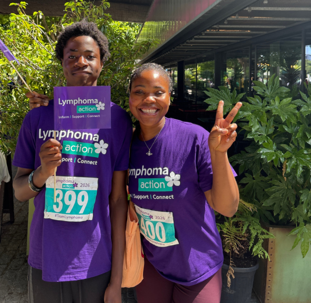 Two Lymphoma Action supporters smiling to camera. Both are wearing Lymphoma Action t shirts and one supporter is waving a branded flag. 