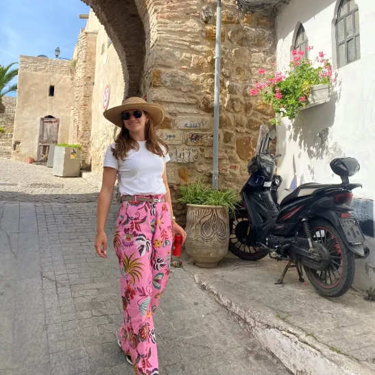 A holiday photo of Katrina. She is walking down a cobbled street wearing a sunhat and sunglasses. A motorbike can be seen in the background. 
