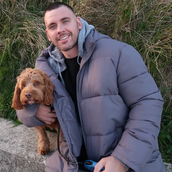 Matthew sitting on a stone wall and smiling to camera, with his arm around his dog. 